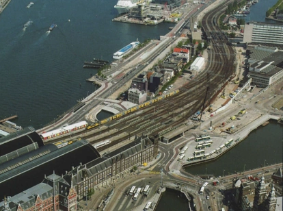 Luchtfoto van de De Ruijterkade in Amsterdam met het Centraal Station, treinsporen, tram- en busstations en het IJ op de achtergrond.